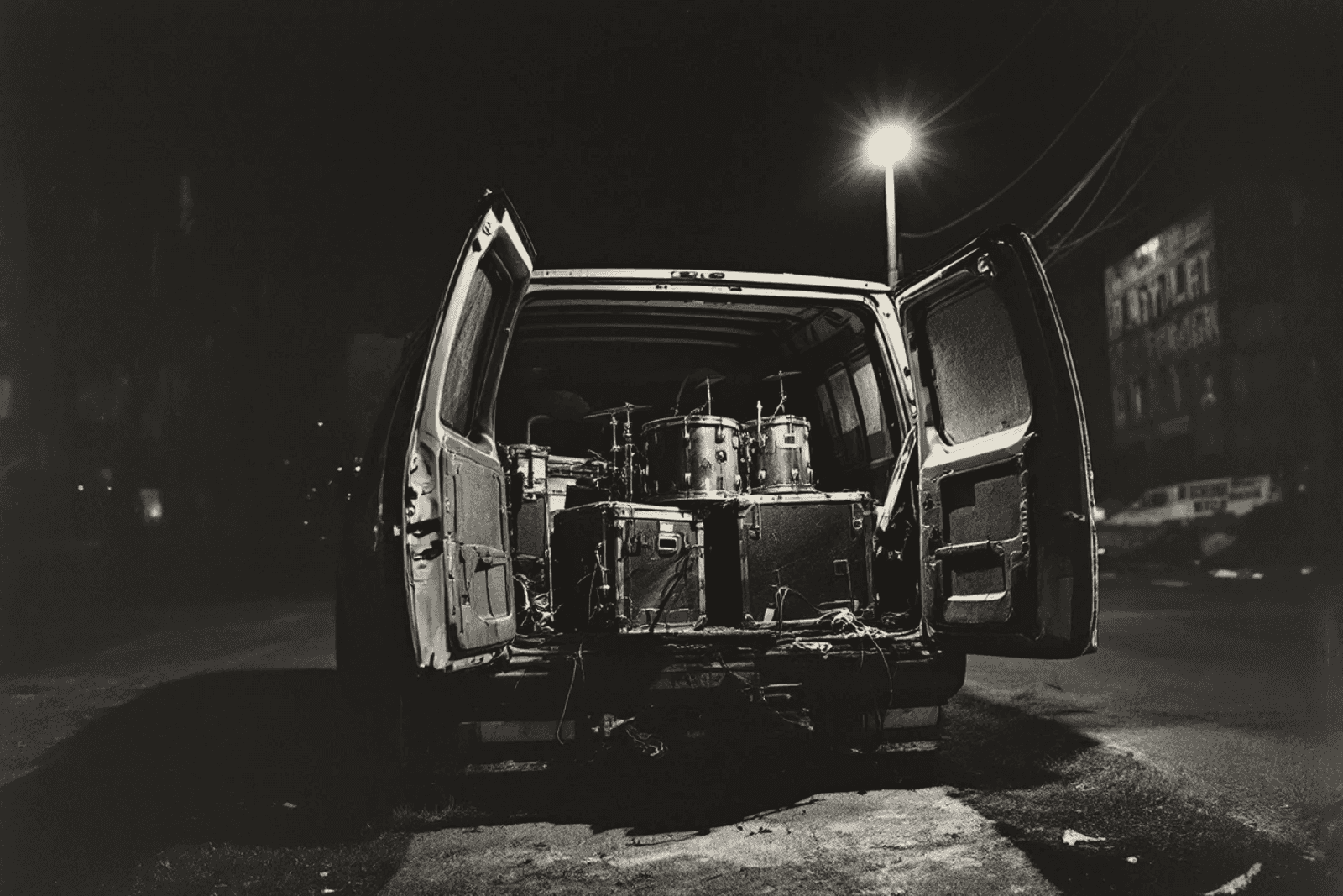 The open back doors of a van at night, a drum kit and flight cases packed inside, a street lamp burning over the empty road behind.