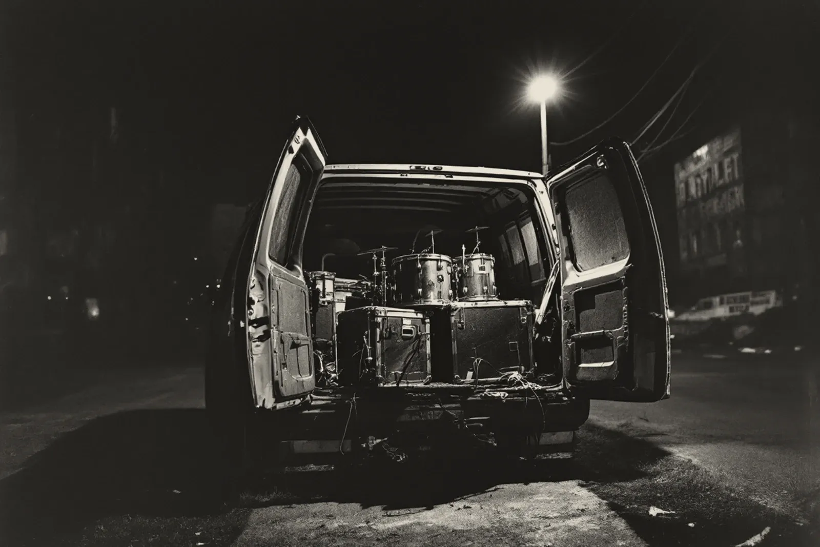 The open back doors of a van at night, a drum kit and flight cases packed inside, a street lamp burning over the empty road behind.