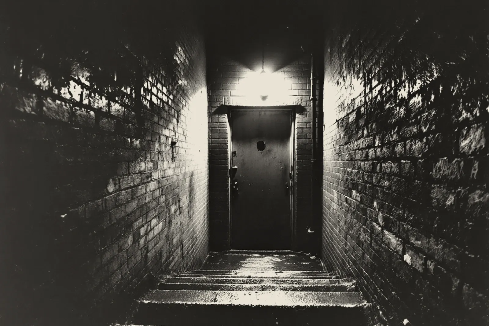 A stairwell going down between bare brick walls to a dented steel door, one bulb burning above it, the steps wet.
